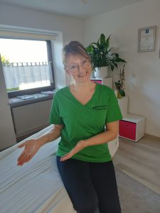 Monika Probst in ihrer Gesundheitswerkstatt. Sie sitzt auf der LIege und deutet einladend auf die Fläche.